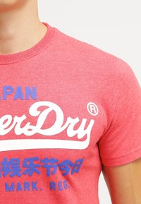 T-shirt rossa in cotone con maniche corte, con grandi scritte bianche e blu e caratteri asiatici sulla parte anteriore. Tessuto liscio.