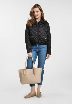 Kobieta ubrana w czarną pikowaną kurtkę i niebieskie dżinsy, trzymająca dużą beżową torebkę, stojąca na tle jasnego, jednolitego tła.