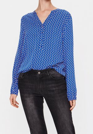 Vrouw draagt een blauwe blouse met lange mouwen met een wit geometrisch patroon en zwarte aansluitende spijkerbroek tegen een witte achtergrond.