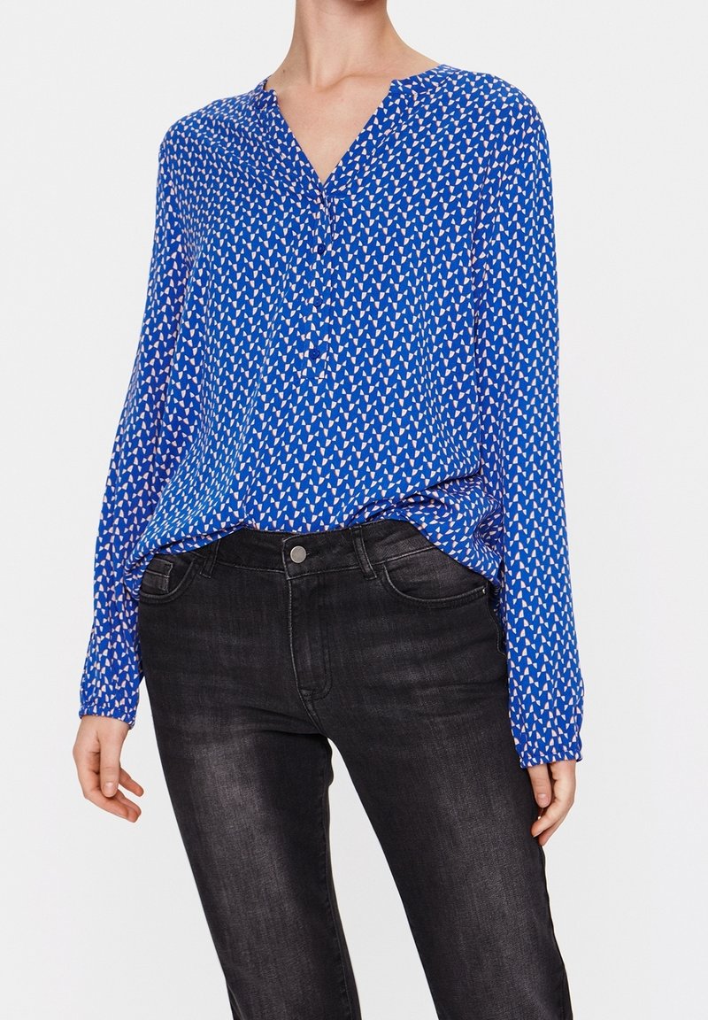Femme portant un chemisier bleu à manches longues avec un motif géométrique blanc et un jean noir ajusté sur fond blanc.