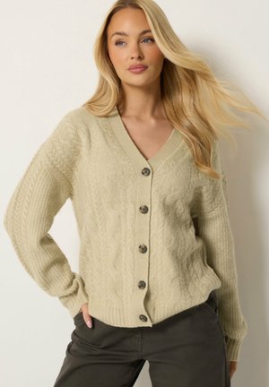 Femme blonde portant un cardigan beige en maille torsadée boutonné et un pantalon foncé, posant avec une main dans la poche sur un fond clair.