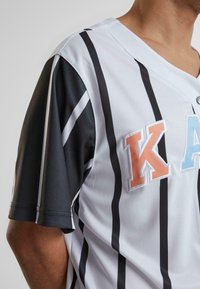 Balta ir juoda sportinė marškinė su trumpomis rankovėmis, su juodu tekstūruotu petimi ir išsiuvinėta „KA“ oranžine ir mėlyna spalvomis.