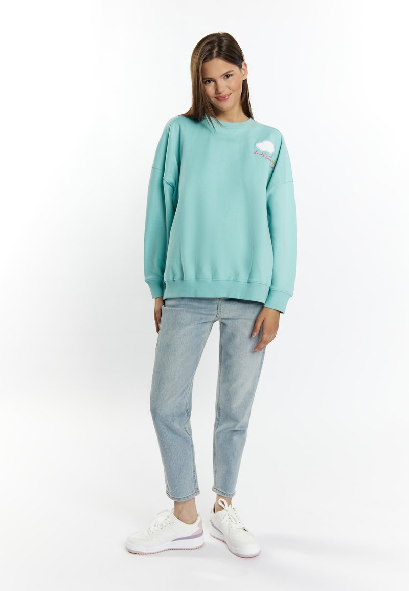 Lichtblauwe sweatshirt met een ronde hals en een losse fit, met een wolkontwerp op de borst. Gepaard met lichte jeans en witte sneakers.