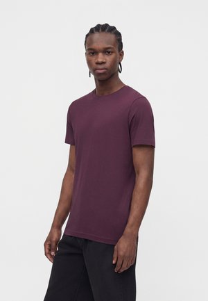Ung mand med flettet hår iført en ensfarvet mørkelilla T-shirt med korte ærmer og sorte bukser, stående mod en hvid baggrund.