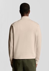 Camicia beige a maniche lunghe con colletto, caratterizzata da una texture liscia. Indossata con pantaloni verde scuro, vista dal retro.