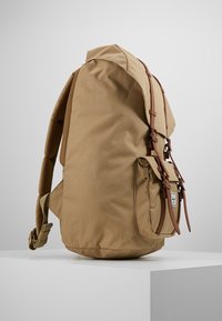 Brązowy plecak z gładkiego płótna, z brązowymi skórzanymi paskami i boczną kieszenią. Zaprojektowany z ergonomicznie ukształtowaną siateczką.