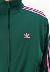 Grønn zip-up jakke laget av glatt stoff, med rosa tre-stripes detaljer på skuldrene og en rosa Adidas-logo på brystet.