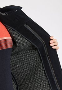Une main tenant un manteau noir ouvert, révélant une doublure intérieure texturée en gris et noir, ainsi qu'une partie d'un pull rayé bleu marine et rouge en dessous.