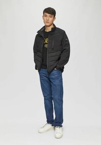 Black padded jacket with zippered pocket, worn over a black graphic t-shirt. Paired with blue jeans and white sneakers. Model stands against a plain background.