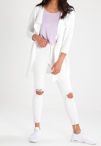 Bílý kardigan s otevřeným předním dílem, vrstvený přes světle fialový top, doplněný o roztrhané bílé džíny a bílé tenisky. Hladká textura, volný design.