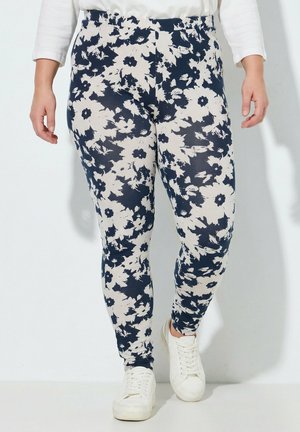 Personne portant des leggings à motifs floraux bleu marine et blanc avec des baskets blanches, debout devant un fond clair uni.