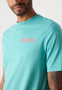 Mand iført en turkis Scotch & Soda t-shirt med korte ærmer og synlig armtatovering mod en ensfarvet baggrund.