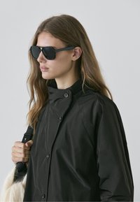 Veste imperméable noire avec col montant, boutons-pression et design élégant ; associée à des lunettes de soleil noires surdimensionnées. Détail du sac à main souple visible.