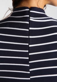 Granatowy materiał w poziome białe paski. Posiada wysoki dekolt oraz zamek błyskawiczny z tyłu na środku do zapinania. Gładkie, teksturowane wykończenie.