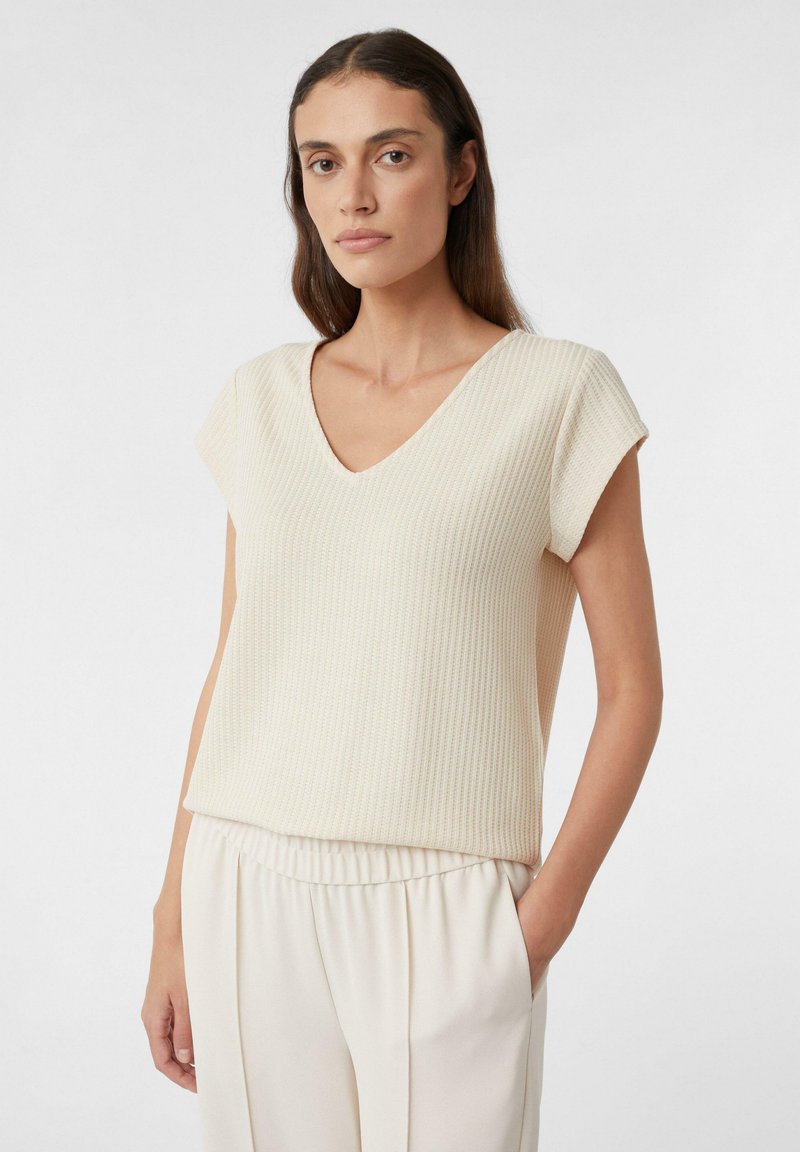 Beige, geribbelde gebreide top met een V-hals en korte mouwen. De textuur is zacht en iets rekbaar, ontworpen voor een ontspannen pasvorm.