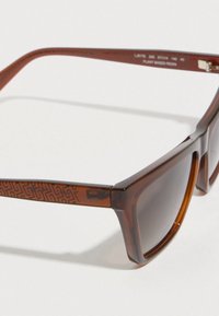 Négyszögletes keretű barna napszemüveg, növényi eredetű gyanta anyagból készült. A barna szárnál geometriai mintázat található, sötét lencsékkel.