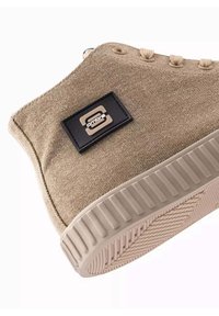 Zapatilla de lona beige con parte superior texturizada, suela de goma y parche de logo negro. Presenta acentos beige contrastantes y un diseño de caña alta.
