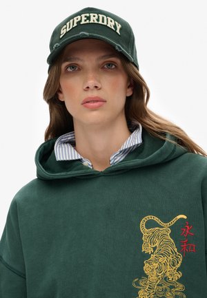 Sudadera verde con capucha, con un gráfico de tigre dorado y texto rojo, superpuesta sobre una camisa de rayas azules y blancas con cuello. Logo de Caps visible.