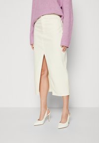 Falda midi de mezclilla color crema con una abertura frontal, combinada con zapatos de tacón blancos. El suéter es de color púrpura claro y presenta un tejido acanalado.