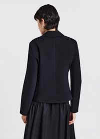 Giacca nera su misura in tessuto strutturato con scollo con colletto e silhouette aderente, abbinata a una gonna nera fluente.