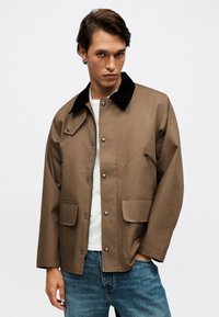 Νέος άντρας που φορά ανοιχτό καφέ μπουφάν με μαύρο γιακά, λευκό πουκάμισο και μπλε τζιν, στέκεται μπροστά σε απλό φωτεινό φόντο.