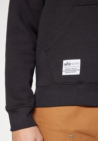 Black sweatshirt with a front pocket and a label patch. The texture is smooth, and it features ribbed cuffs and hem. Worn over brown pants.