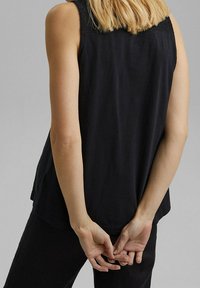 Svart ärmlös topp med en texturerad spetspanel på baksidan, med en lös siluett och mjukt tyg. Händerna är knäppta bakom.