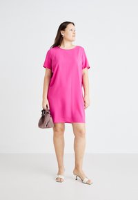 Vestido rosa de mangas curtas com um corte descontraído, textura suave e decote redondo. Combinado com uma pequena bolsa castanha franzida e sapatos de salto aberto.