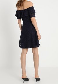 Žena v námořnickém modrém šatech bez ramínek z krajky a černých vysokých podpatcích, stojící zády k jednoduchému bílému pozadí.