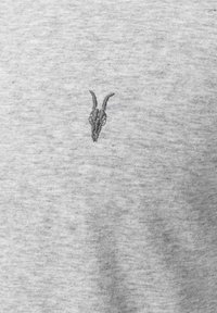 Broderie d'un petit crâne de chèvre sombre avec des cornes courbées sur un tissu gris clair, centré horizontalement.