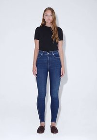 Blå jeans med hög midja i smal passform, med en klassisk design med fem fickor och subtila veckdetaljer. Kombinerade med en svart kortärmad topp.