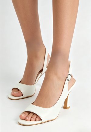 Pieds portant des talons hauts blancs à bout ouvert et bride arrière avec boucles, présentés sur un fond blanc uni.