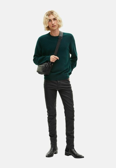 Młoda osoba o blond włosach, ubrana w ciemnozielony sweter, czarne skórzane spodnie, czarne buty i nosząca czarną torbę na ramię.