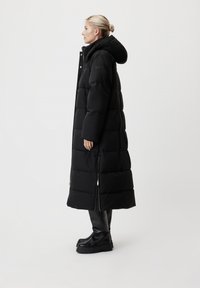 Femme de profil portant un long manteau matelassé noir avec capuche, des bottes noires épaisses, se tenant contre un fond blanc uni.