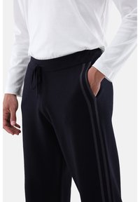 Mann trägt schwarze Sweatpants mit seitlichen Streifen und ein weißes Langarmshirt, Hand in der Tasche, Nahaufnahme auf Taille und Hüften.