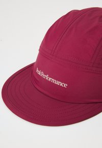 Marron kasket lavet af glat stof med en buet skygge. Har syningsdetaljer og "PeakPerformance" logo i hvidt på frontpanelet.