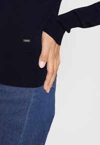 Laivaston sininen neulepusero, jossa on resorit hihansuissa ja helmassa, pieni metallinen logovivahde, yhdistetty farkkuihin. Lähikuva alaosasta.