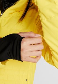 Gelbe Pufferjacke mit einer gesteppten Textur und einem schwarzen inneren Ärmel. Handreißverschluss-Seitentasche mit gelbem Zipper.