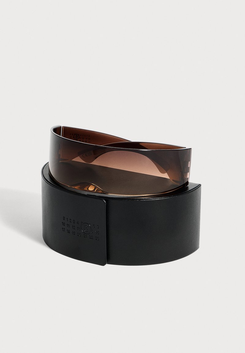 Ceinture en cuir noir enroulée à côté d'une visière en plastique brun translucide incurvée sans fixations visibles sur un fond clair uni.
