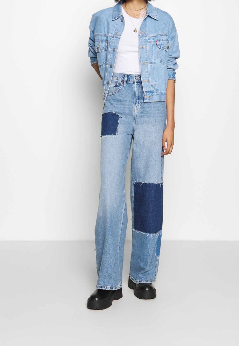 Vaaleansininen farkkutakki, jossa on muotoiltu malli, yhdistettyinä leveälahkeisiin farkkuihin, joissa on tummansinistä tilkkutöitä ja repsottavat reunat. Mustat platform-kengät.