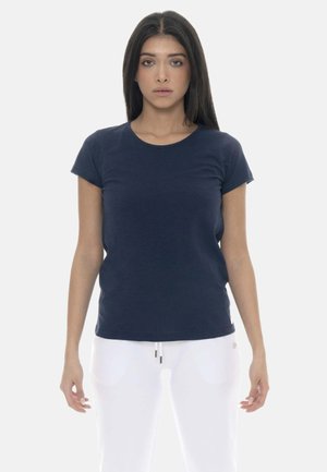 Giovane donna con lunghi capelli scuri che indossa una maglietta blu navy a maniche corte e pantaloni bianchi, in piedi davanti a uno sfondo chiaro e semplice.