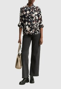 Blusa negra con estampado floral y mangas enrolladas, combinada con jeans de pierna ancha de tiro alto en negro y botines gruesos negros, sosteniendo un bolso beige.