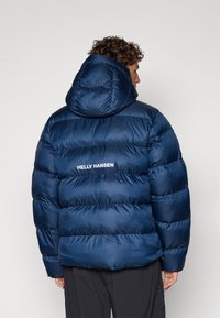 Doudoune bleu marine avec capuche ; design matelassé et texture brillante ; présente le logo blanc "HELly HANSEN" au dos.