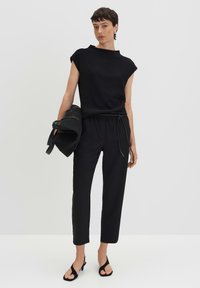 Schwarzes kurzärmliges, strukturiertes Oberteil, kombiniert mit enganliegenden schwarzen Hosen mit einstellbarem Kordelzug. Das Model hält eine schwarze Tasche und steht in Sandalen.