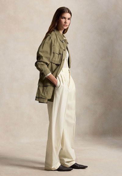 Γυναίκα που στέκεται πλάγια φορώντας ένα χακί μπουφάν, κρεμ παντελόνες με φαρδιά πόδια και μαύρα μυτερά παπούτσια σε ουδέτερο φόντο.