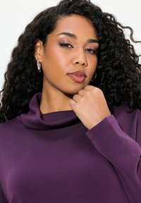 Maglia viola con scollo a cowl realizzata in tessuto morbido, dotata di maniche lunghe e una silhouette aderente. Presenta un dettaglio testurizzato attorno al collo.