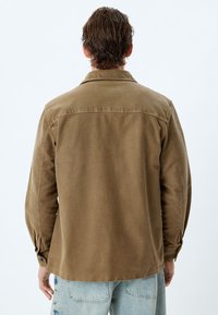 Camisa marrón de botones, de manga larga y corte relajado, hecha de un tejido resistente. Presenta un collar y puños clásicos. Vista trasera de estilo casual.