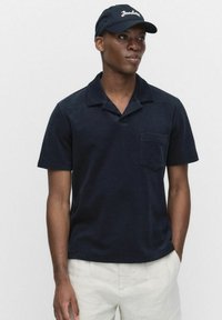 Marineblauw poloshirt met korte mouwen en kraag, zakje op de linkerborst, zachte textuur. Draagt een bijpassende donkere pet.