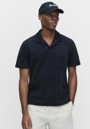 Navy short-sleeve polo shirt with a collar, pocket on the left chest, soft texture. Worn with a matching dark baseball cap.