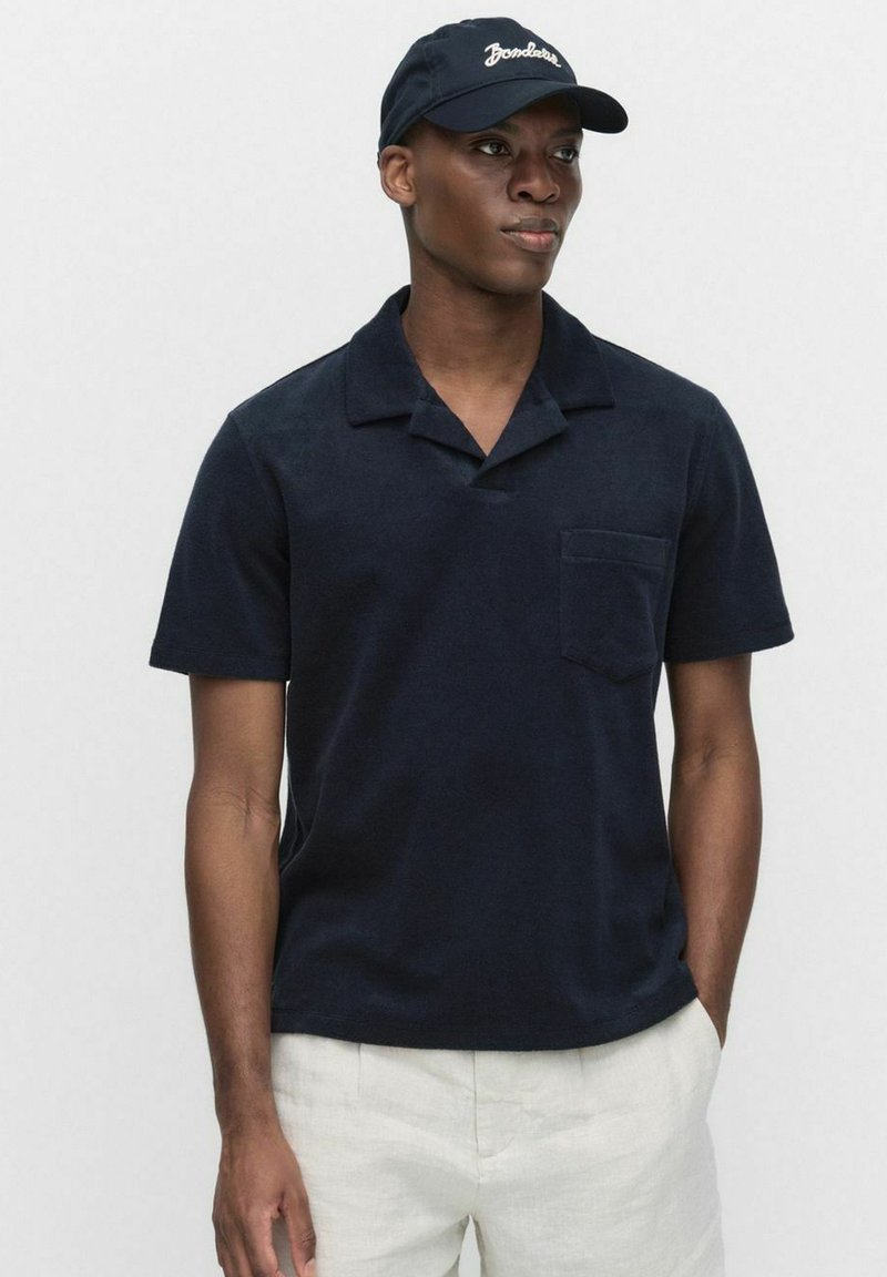 Marineblauw poloshirt met korte mouwen en kraag, zakje op de linkerborst, zachte textuur. Draagt een bijpassende donkere pet.